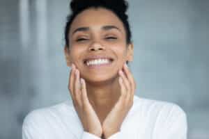 Young black woman happily touching her face after getting skin rejuvenation treatment.